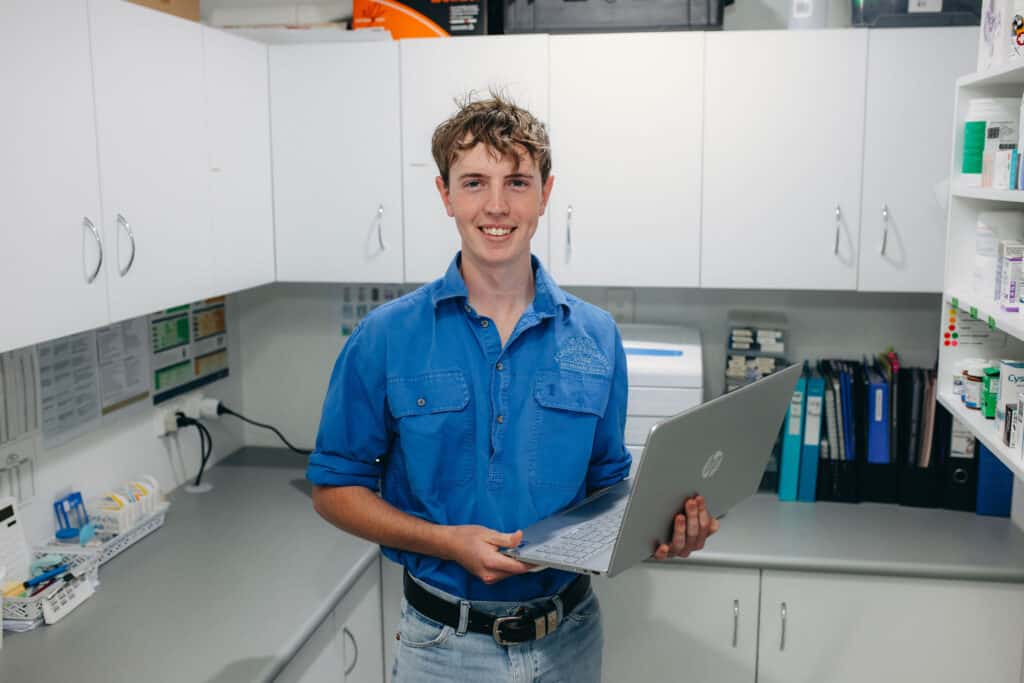 A young man in a pharmacy holding a laptop, possibly researching medication or seeking medical advice.
